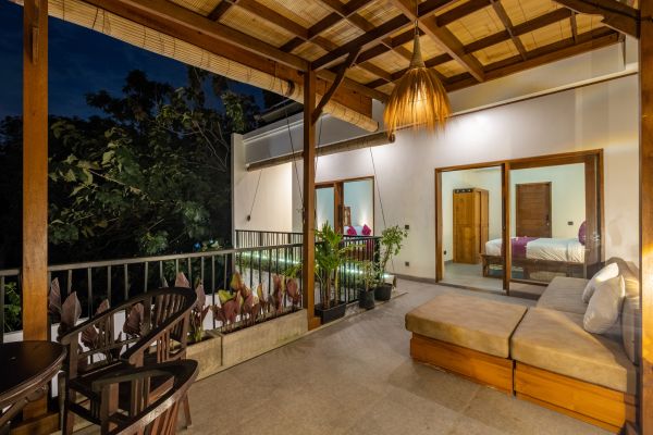 Cozy tropical balcony at night with wooden veranda, hanging rattan lights, lounge chairs, potted plants, and sliding doors to a bedroom overlooking lush greenery.