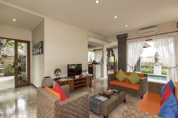 Modern Balinese villa living room with rattan sofas in orange, TV console, glass coffee table, and serene pool garden view through sheer-curtained doors. - Bali Villas