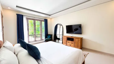 Spacious modern tropical bedroom with king bed, blue velvet armchair, wooden TV cabinet, and arched windows with sheer blue curtains.