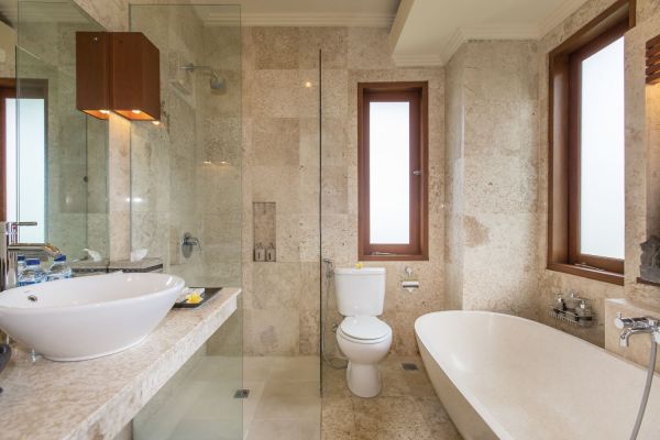 Modern luxury bathroom with beige stone tiles, freestanding white bathtub, glass shower enclosure, and wooden accents. - Bali Villas