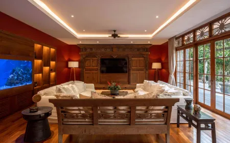 Luxurious Balinese living room with carved wooden furniture, white sectional sofas, aquarium, large TV, and sliding doors to tropical deck. - Bali Villas