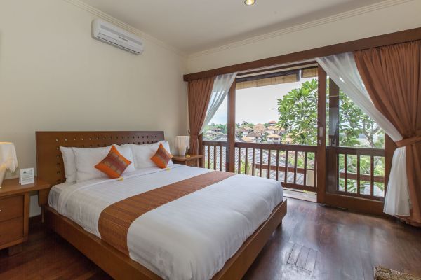 Spacious Balinese-style bedroom with wooden king bed, open balcony doors, and scenic view of tropical trees and village rooftops.