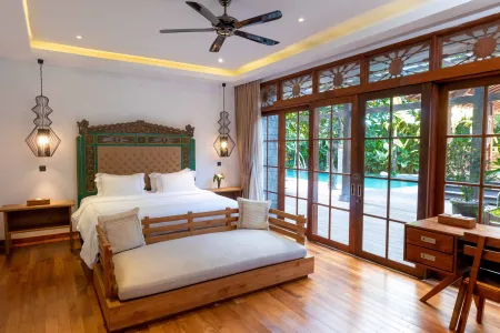 Luxurious Balinese bedroom with carved wooden bed, daybed, and tropical pool view through floor-to-ceiling sliding doors.