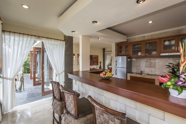 Modern tropical kitchen with wooden bar counter, stools, fresh fruits and flowers, open folding doors to patio, and Balinese decor. - Bali Villas
