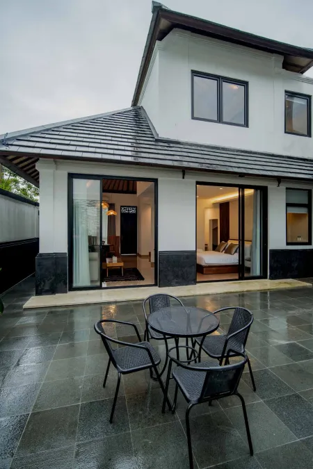 Modern Indonesian-style house exterior with tiled roof, rainy slate patio, metal furniture, and open glass doors to a bedroom.