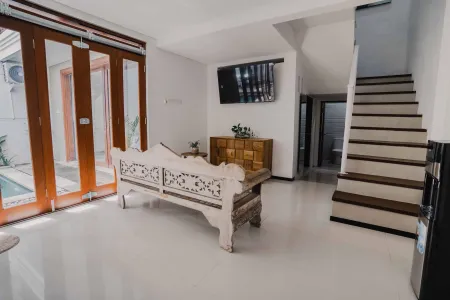 Serene modern Indonesian living room with carved wooden bench, open bifold doors to pool courtyard, wall TV, and staircase. - Bali Villas