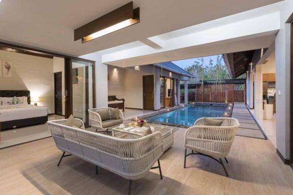 Luxurious open-plan Balinese villa bedroom with white wicker seating and infinity pool view.