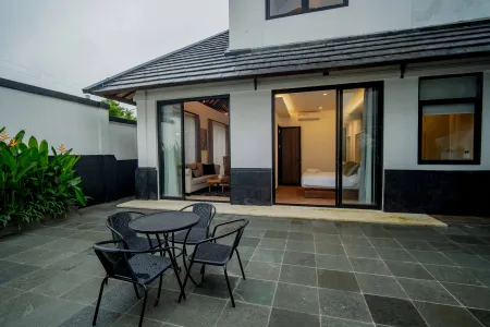 Modern tropical villa patio with slate tiles, black metal table and chairs, open sliding glass doors to a cozy bedroom, surrounded by lush plants.