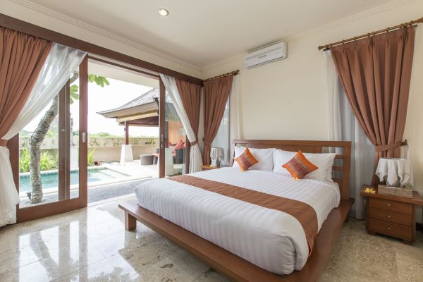Spacious Balinese bedroom with wooden king bed, orange-accented linens, and sliding doors to a private outdoor pool. - Bali Villas