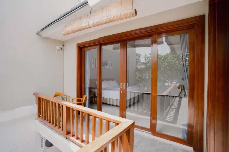 Spacious wooden balcony with sliding doors to a bedroom, bamboo shades overhead, and a small chair against a white wall.