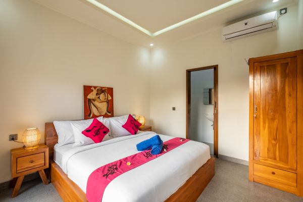 Cozy Balinese villa bedroom with white king bed, pink pillows, wooden furniture, and tropical wall art.