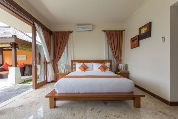 Spacious Balinese bedroom with wooden platform bed, white linens, orange accents, and open sliding doors to outdoor pool patio.