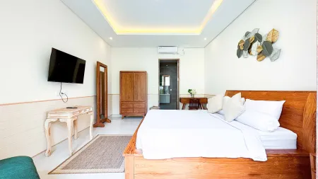 Spacious Balinese-style bedroom with wooden king bed, wall-mounted TV, ornate wardrobe, and tropical wall art.