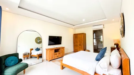 Spacious tropical bedroom with wooden king bed, wall-mounted TV, arched mirror, and green accent chair.