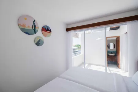 Bright minimalist bedroom with white bed, pastel desert landscape wall art, sliding doors to sunny balcony, and en-suite bathroom.