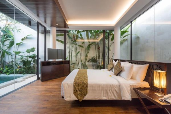 Luxurious Bali villa bedroom with king bed, tropical plants, and indoor pool view through glass walls.