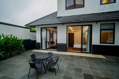 Modern tropical villa exterior with open sliding glass doors to a bedroom, featuring a patio table and chairs amid lush greenery.