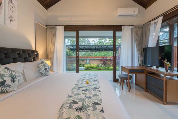 Serene Balinese villa bedroom with plush king bed, wooden accents, and lush garden view through open sliding doors.