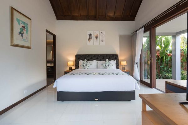 Spacious Balinese-style bedroom with king bed, tropical garden view through open sliding doors, wooden accents, and feather wall art.