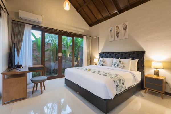 Cozy Balinese villa bedroom featuring a king-sized bed with white floral bedding, sliding glass doors to lush garden, wooden furniture, and soft ambient lighting.