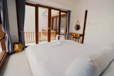 Bright, airy tropical bedroom with white king bed, swan-shaped towel, wooden desk and mirror, and open folding doors to balcony.