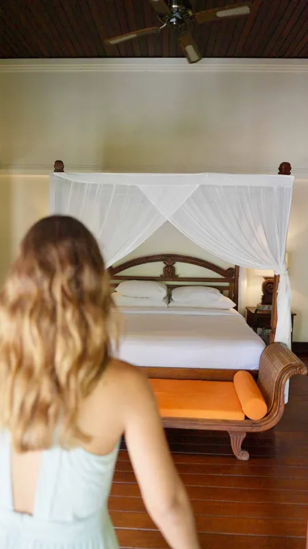 Woman with long wavy hair stands in a luxurious tropical bedroom, facing a white canopy bed with mosquito netting and an orange chaise lounge on wooden floors.