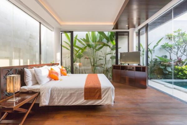 Luxurious tropical bedroom with king bed, orange accents, potted plants, and glass walls opening to an infinity pool.