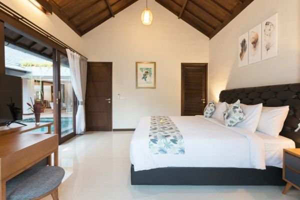 Luxurious Balinese villa bedroom with king bed, wooden beamed ceiling, and poolside view through sliding doors.