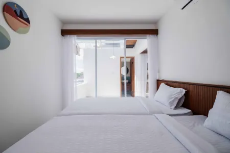 Minimalist white bedroom with two neatly made twin beds, wooden headboards, abstract wall art, and sheer-curtained sliding doors to outdoor greenery.