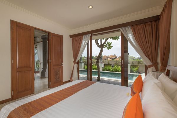 Spacious Bali villa bedroom with king bed, orange accents, and sliding doors opening to a private pool and tropical garden view.