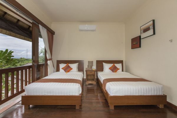 Cozy Balinese-style twin bedroom with wooden beds, orange accents, and balcony view of tropical greenery.