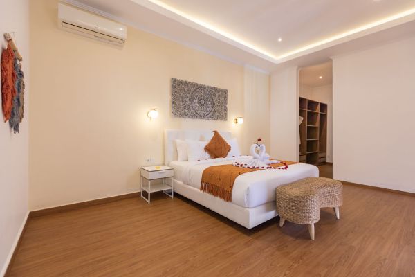 Elegant tropical hotel bedroom with white king bed, orange bedding, rose petals, swan towel, and woven wall art on beige walls.