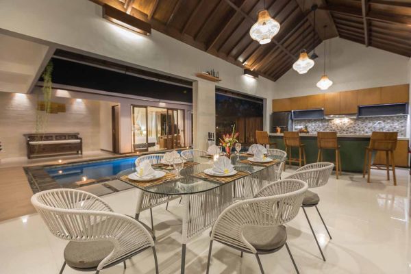 Elegant open-plan dining room in a luxury Balinese villa, featuring a glass-top table with rattan chairs, tropical centerpiece, sleek kitchen, and poolside views. - Bali Villas