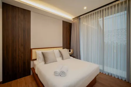 Modern bedroom interior with wooden bed, white duvet, gray pillows, and sheer curtains on large windows.