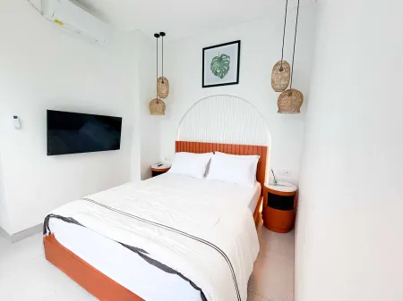 Cozy modern bedroom with white bed on orange base, wall-mounted TV, rattan pendant lights, and tropical leaf art.