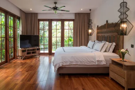 Spacious tropical bedroom with wooden king bed, large glass doors to lush garden, ceiling fan, and flat-screen TV.