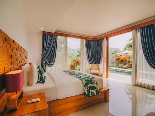 Luxurious Bali villa bedroom with wooden headboard, tropical leaf-patterned king bed, sheer blue curtains, and infinity pool views through floor-to-ceiling windows.