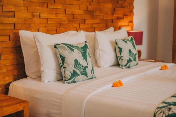 Cozy Balinese bedroom with teak wood headboard, crisp white linens, green leaf-patterned pillows, and orange frangipani petals on the bed.