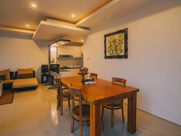 Modern open-plan living and dining space with wooden table, chairs, fruit bowl, L-shaped sofa, kitchen counters, and floral wall art. - Bali Villas