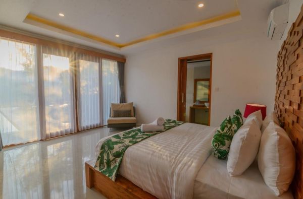 Spacious tropical bedroom with wooden bed, green leafy bedding, large sheer-curtained windows, and warm sunlight.