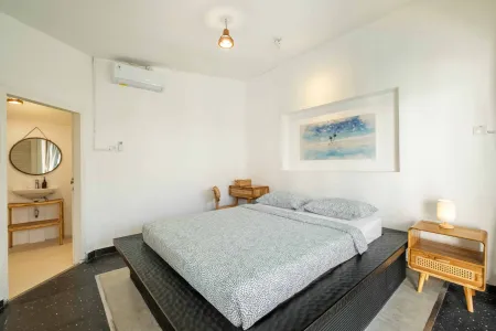 Modern minimalist bedroom with platform bed, rattan furniture, abstract ocean art, and open bathroom in white tones.