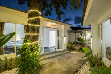 Tropical Bali villa at dusk: fairy lights wrap a large tree beside open glass doors to a white bedroom, lush plants line a stone path.