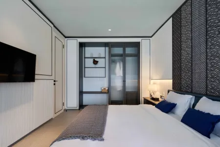 Modern minimalist hotel bedroom with white bed, blue accent pillows, wall-mounted TV, and bamboo wall panels.