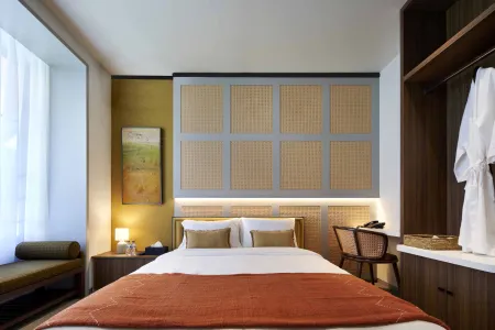 Modern hotel bedroom with bamboo wall panels, queen bed topped with orange throw and pillows, wooden desk, rattan chair, and sheer-curtained window.