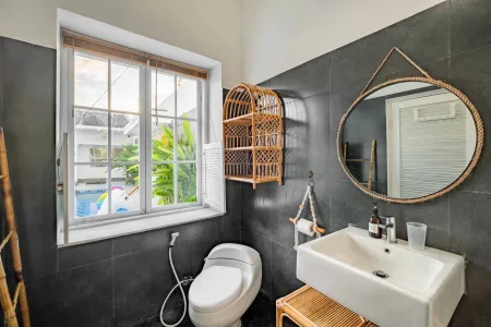 Modern tropical bathroom with dark tiled walls, rattan shelf and bamboo accents, oval rope mirror, white sink, bidet toilet, and window view of poolside greenery. - Bali Villas