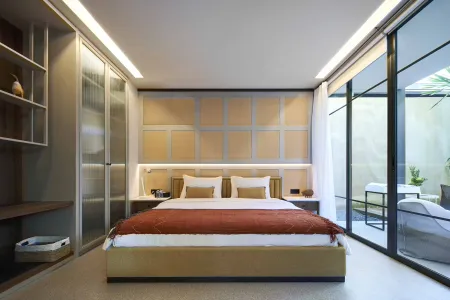 Modern minimalist bedroom with plush beige upholstered bed, rust throw blanket, built-in wardrobes, and floor-to-ceiling glass doors opening to a lush outdoor patio.