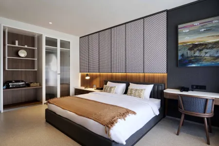 Modern minimalist hotel bedroom featuring a king bed with orange throw, wooden headboard, mesh partition, built-in wardrobe, desk, and abstract artwork. - Bali Villas