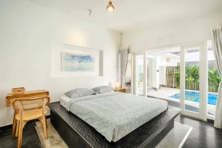 Modern Balinese bedroom with platform bed, wooden desk, abstract wall art, and sliding doors to private pool.
