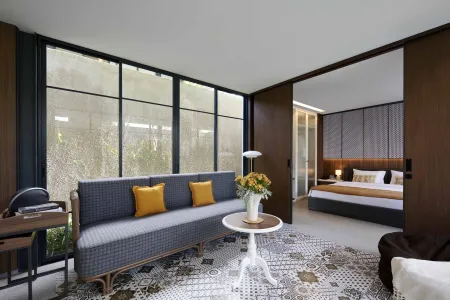 Modern hotel suite interior with gray sofa, yellow pillows, flower vase on white table, large windows, sliding wooden doors, and adjacent bedroom. - Bali Villas
