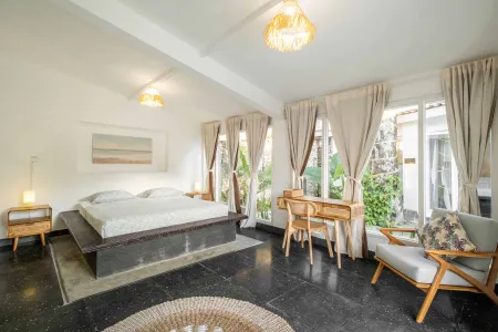 Serene tropical bedroom with platform bed, wooden desk and armchair, large curtained windows overlooking lush garden, pendant lights, and woven rug on black tile floor.
